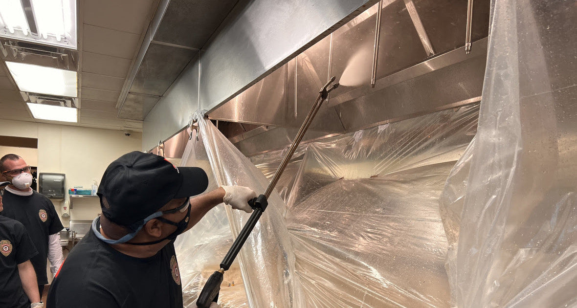 Technician cleaning a commercial kitchen exhaust hood with a pressure washer while surrounding equipment is protected with plastic sheeting during a professional kitchen exhaust hood cleaning service to help maintain NFPA 96 fire safety compliance.